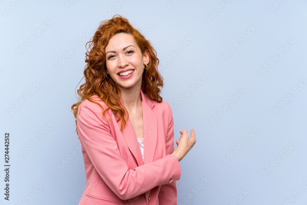 Redhead woman in suit over isolated blue wall pointing back Redhead woman in suit over isolated blue wall pointing back