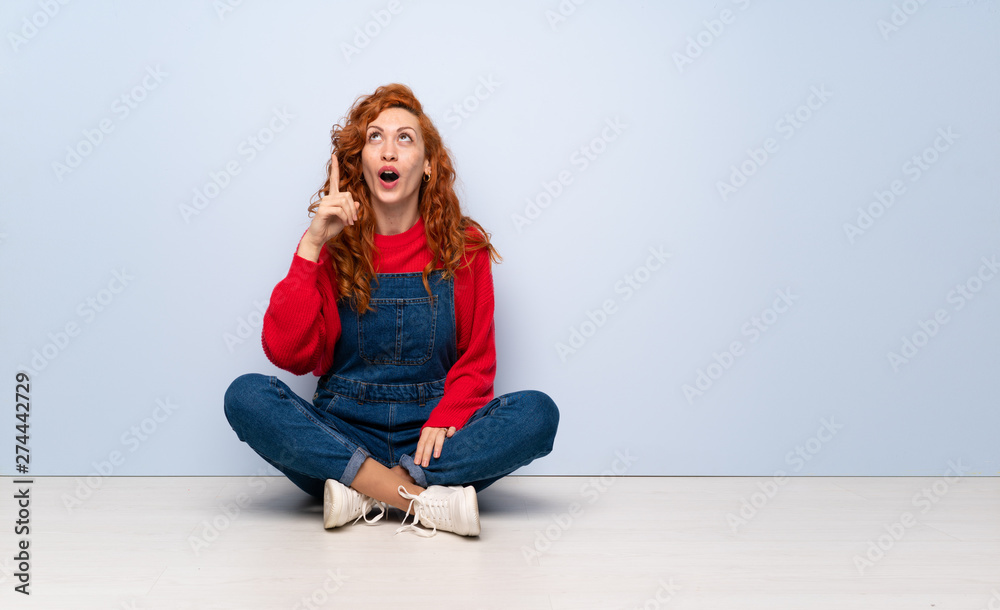 Redhead woman with overalls sitting on the floor pointing up and surprised Redhead woman with overalls sitting on the floor pointing up and surprised