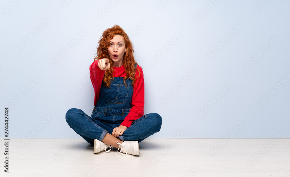 Redhead woman with overalls sitting on the floor surprised and pointing front Redhead woman with overalls sitting on the floor surprised and pointing front