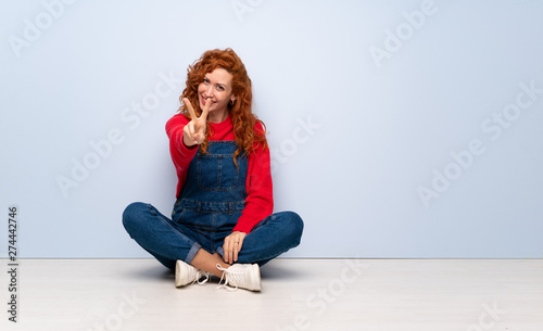 Redhead woman with overalls sitting on the floor smiling and showing victory sign
