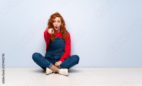 Redhead woman with overalls sitting on the floor surprised and pointing front