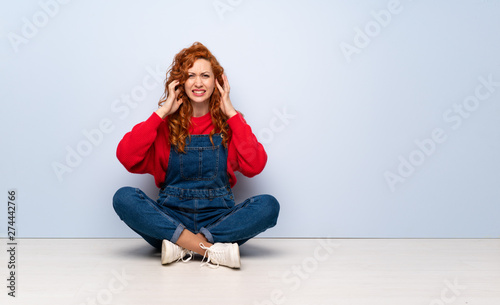 Redhead woman with overalls sitting on the floor frustrated and covering ears