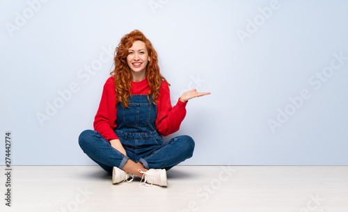 Redhead woman with overalls sitting on the floor holding copyspace imaginary on the palm to insert an ad