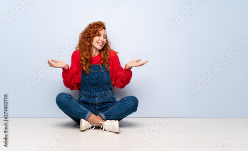 Redhead woman with overalls sitting on the floor holding copyspace with two hands