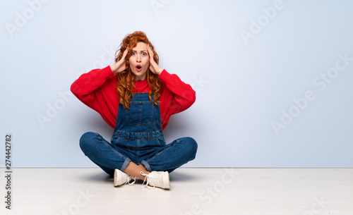 Redhead woman with overalls sitting on the floor with surprise expression