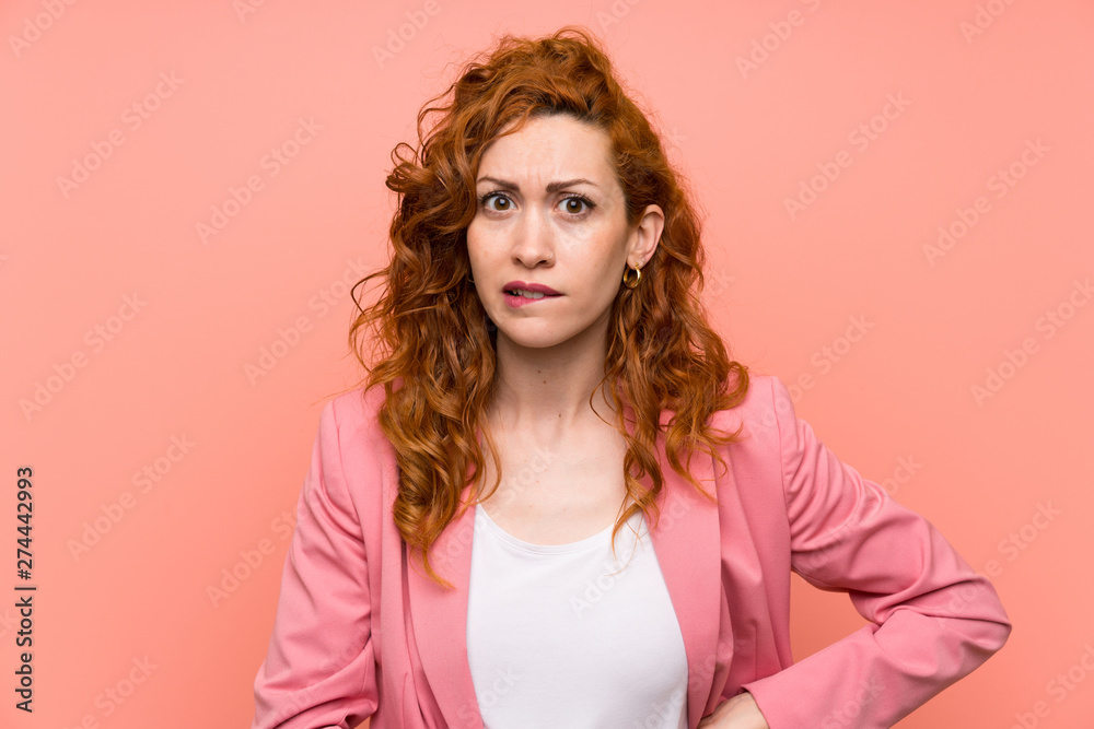 Redhead woman in suit over isolated pink wall having doubts and with confuse face expression Redhead woman in suit over isolated pink wall having doubts and with confuse face expression