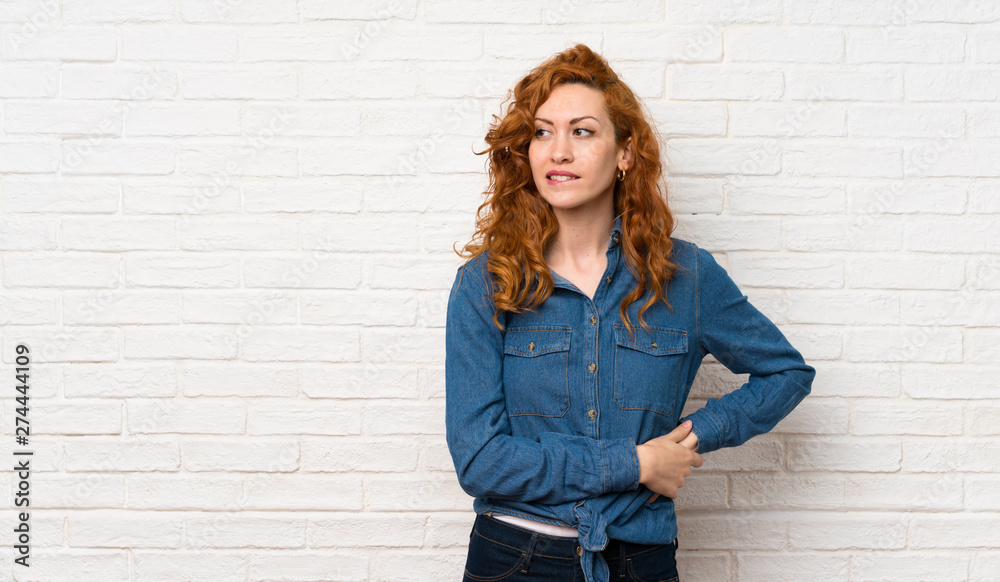 Redhead woman over white brick wall with confuse face expression Redhead woman over white brick wall with confuse face expression