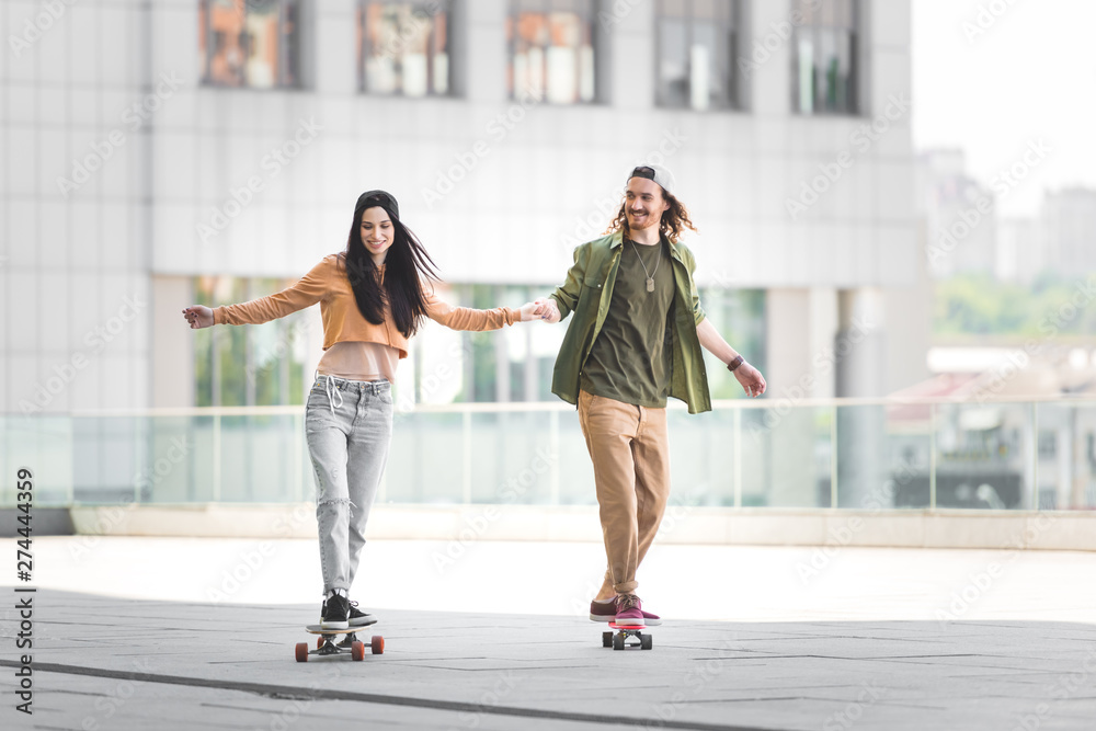 happy man holding hands with woman, riding on skateboard in city