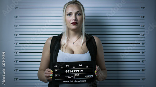 In a Police Station Arrested Woman Gets Front-View Mug Shot. She Wears Saucy Clothes, Has Heavy Makeup and Holds Placard. Height Chart in the Background. Shot with Dark Cold Lights and Vignette Filter