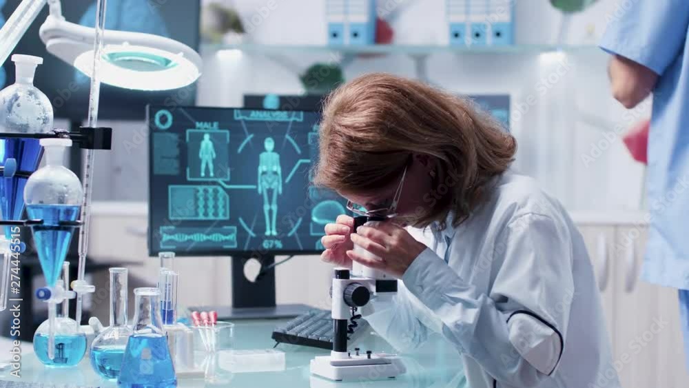 Female scientist uses a microscope to analyze some samples. In the background - an active team is working and a monitor displays an advanced scan system Female scientist uses a microscope to analyze some samples. In the background - an active team is working and a monitor displays an advanced scan system