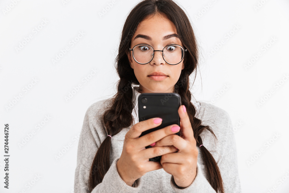 Photo closeup of puzzled teenage girl wearing eyeglasses and school uniform wondering while holding smartphone Photo closeup of puzzled teenage girl wearing eyeglasses and school uniform wondering while holding smartphone