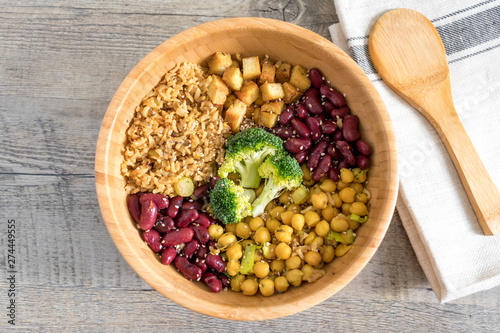 Vegan buddha bowl with stir fry tofu, brown rice, broccoli, red kidney beans, cooked chickpeas, seeds and vegetables. Flat lay on wooden surface with kitchen towel and ladle.