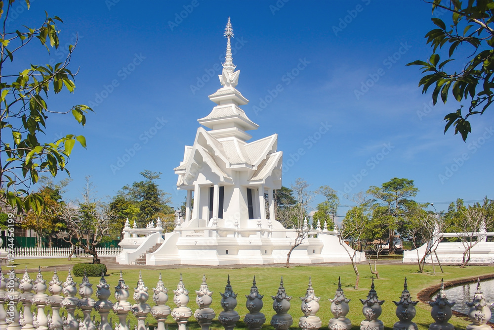 Buddhist building with tiered roof in Wat Rong Khun or White Temple surrounded by trees and balusters with blue sky