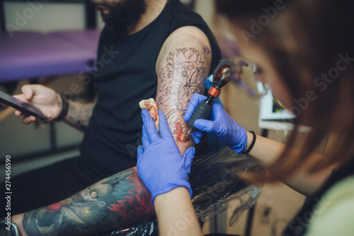 tattooist woman makes a tattoo in her studio to a young man while consulting his smartphone