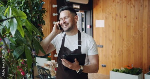 Smiling guy florist in apron is speaking on mobile phone and holding modern tablet at work communicationg to customers smiling. People and conversation concept. photo