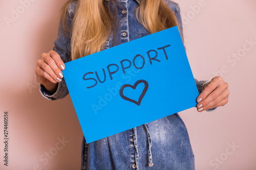 Woman holding paper with word support and heart shape while standing in front of the wall.