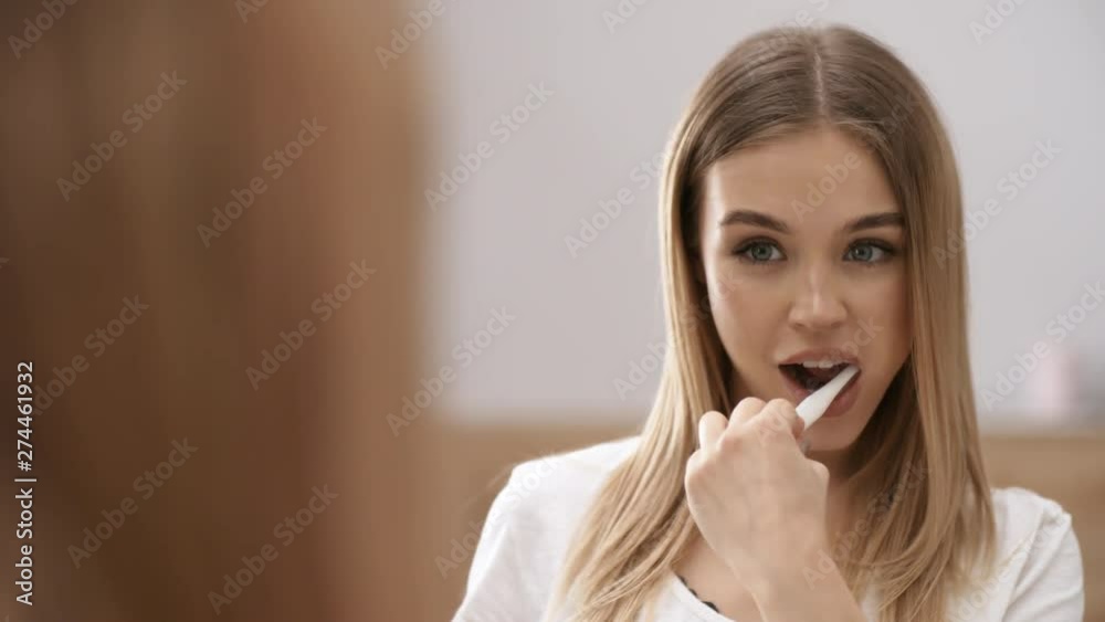 Chest-up mirror shot of gorgeous young Caucasian woman brushing teeth in bathroom and looking at reflection in mirror with delight, smiling to herself, turning her head and striking poses Chest-up mirror shot of gorgeous young Caucasian woman brushing teeth in bathroom and looking at reflection in mirror with delight, smiling to herself, turning her head and striking poses