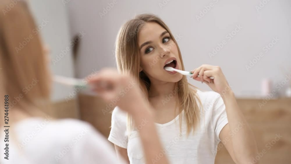 Chest-up mirror shot of glamorous young Caucasian woman with loose long blond hair, wearing white t-shirt, brushing her teeth in front of bathroom mirror, looking straight and admiring her reflection Chest-up mirror shot of glamorous young Caucasian woman with loose long blond hair, wearing white t-shirt, brushing her teeth in front of bathroom mirror, looking straight and admiring her reflection
