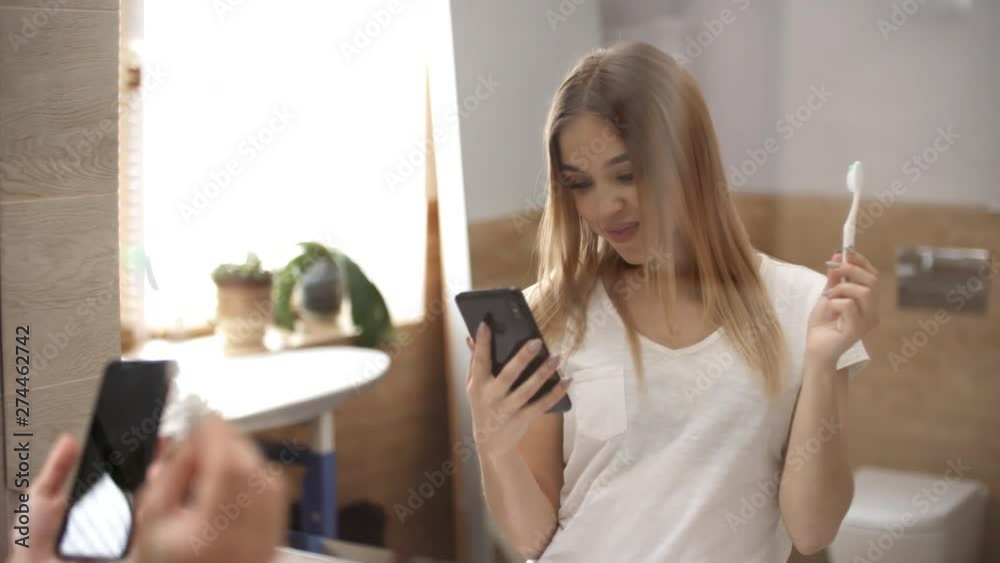 Waist-up mirror shot of beautiful 20-something Caucasian woman brushing her teeth in morning in front of bathroom mirror and checking messages and emails on her mobile phone with excited smile