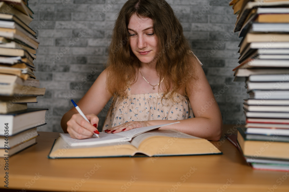 Tired female student reading among books. Pensive young woman sitting at table with pile of book and reading on background of grey wall Tired female student reading among books. Pensive young woman sitting at table with pile of book and reading on background of grey wall