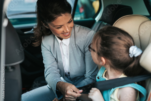 Young businesswoman helping her daughter to fasten seatbelts in the car while girl is sitting on a safety child car seat.
