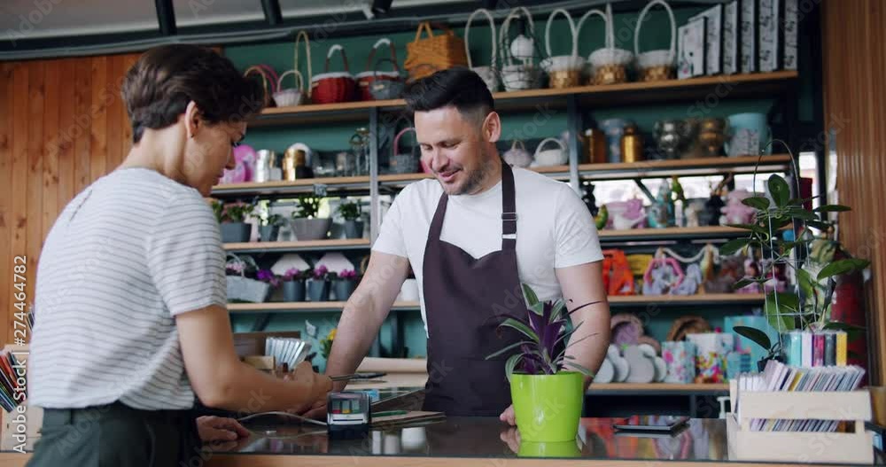 Female customer pretty girl is making payment with smartphone in flower store buying green houseplant talking to salesman smiling. Small business and finance concept.