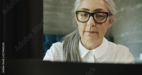 Aged beautiful female in eyeglasses typing on laptop at home, computer literacy for retirees