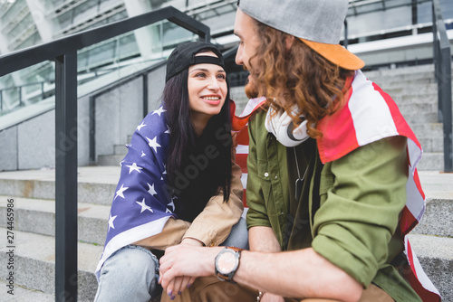 selective focus of couple with american flag on shoulders sitting on stairs, looking at each other
