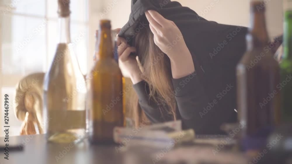 Young woman and the man sitting at the table near empty alcohol bottles. Intoxicated girl covering head with the hood. Unhealthy lifestyle. Troubled teens. Drug addiction. Slow motion.