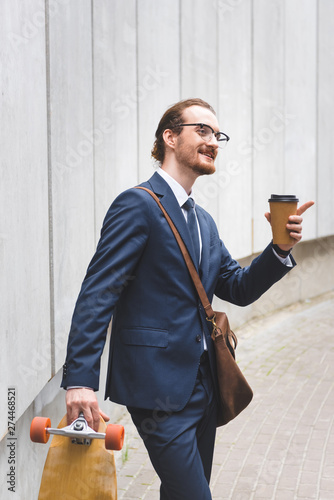 happy businessman in formal wear holding skateboard with paper cup in hand and pointing finger away