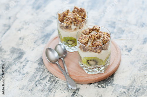 Dessert granola, Greek yogurt, kiwi and banana in two glass cups with spoons on wooden round board, gray concrete, close-up. Fitness, figure, body and healthy food