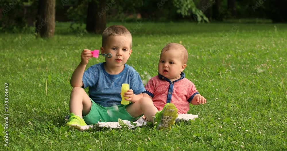Happy little brother playing and hugging his sibling baby sitting on grass in a park. brothers play with soap bubbles concept of love and parenting. Happy little brother playing and hugging his sibling baby sitting on grass in a park. brothers play with soap bubbles concept of love and parenting.