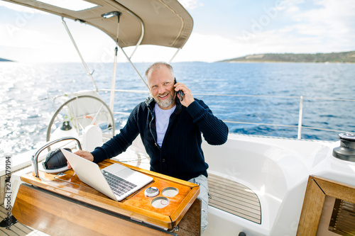 Digitaler Nomade mit Laptop telefoniert verschmitzt lächelnd auf einem Segelboot