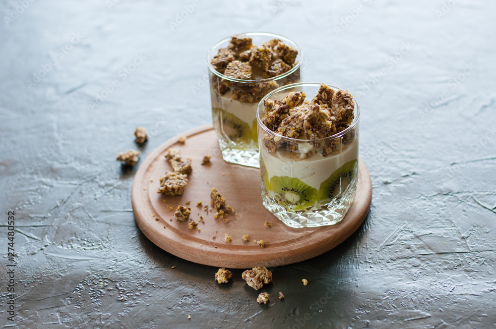 Greek yogurt, granola, banana and kiwi. Desserts in glass cups with spoons on a wooden round shelf, dark background, side view. Concept body and healthy food Greek yogurt, granola, banana and kiwi. Desserts in glass cups with spoons on a wooden round shelf, dark background, side view. Concept body and healthy food