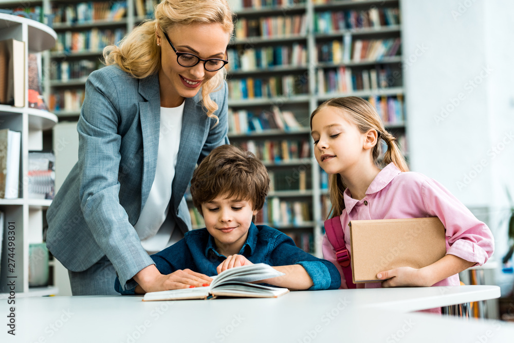 cheerful teacher in glasses standing near cute kids reading book in library cheerful teacher in glasses standing near cute kids reading book in library