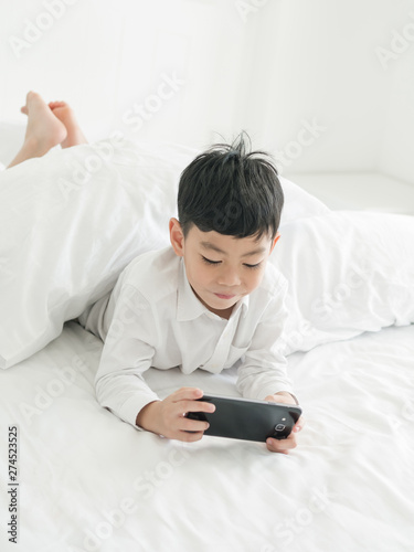 Cute little asian kid focused on smartphone while lying on the bed. Boy playing games on smartphone. Social and technology concept
