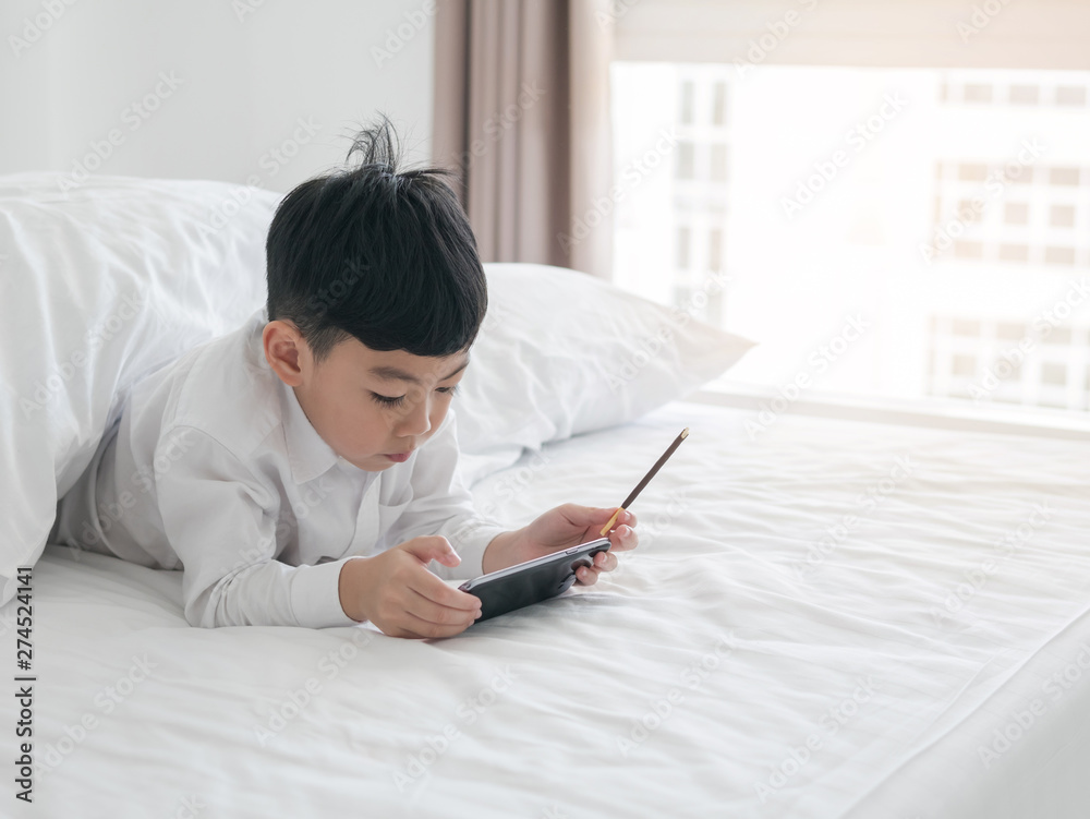 Cute little asian kid focused on smartphone while lying on the bed. Boy playing games on smartphone. Social and technology concept