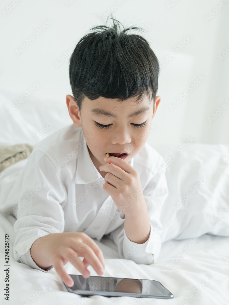 Cute little asian kid focused on smartphone while lying on the bed. Boy playing games on smartphone. Social and technology concept Cute little asian kid focused on smartphone while lying on the bed. Boy playing games on smartphone. Social and technology concept
