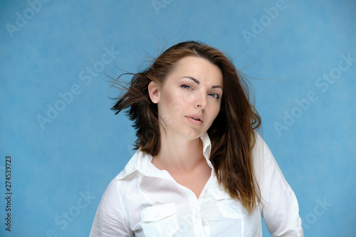 A close-up portrait of a pretty girl, a brunette woman, 30 years old, against a blue background in a white shirt with dark hair and excellent skin. She shows emotions, smiles, wonders. © Вячеслав Чичаев