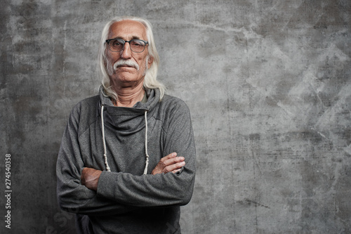 Old Caucasian gray-haired cool grandfather with glasses incredulously looks into the camera with crossed arms and serious face with copy space in studio photo