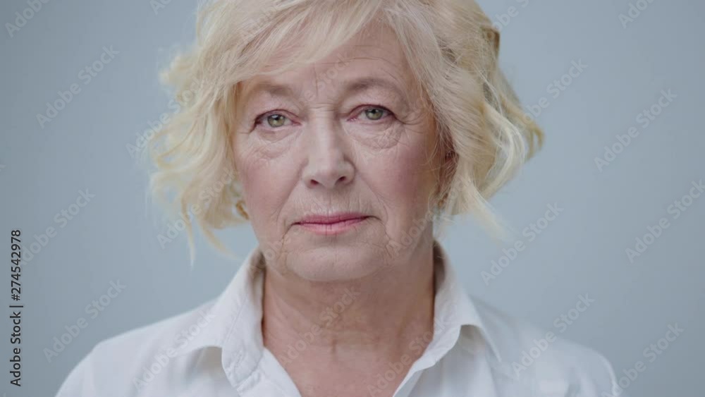 Portrait of nice aged blonde model with green eyes isolated on grey background. Beautiful mature woman with serious expression in white shirt looking at camera.