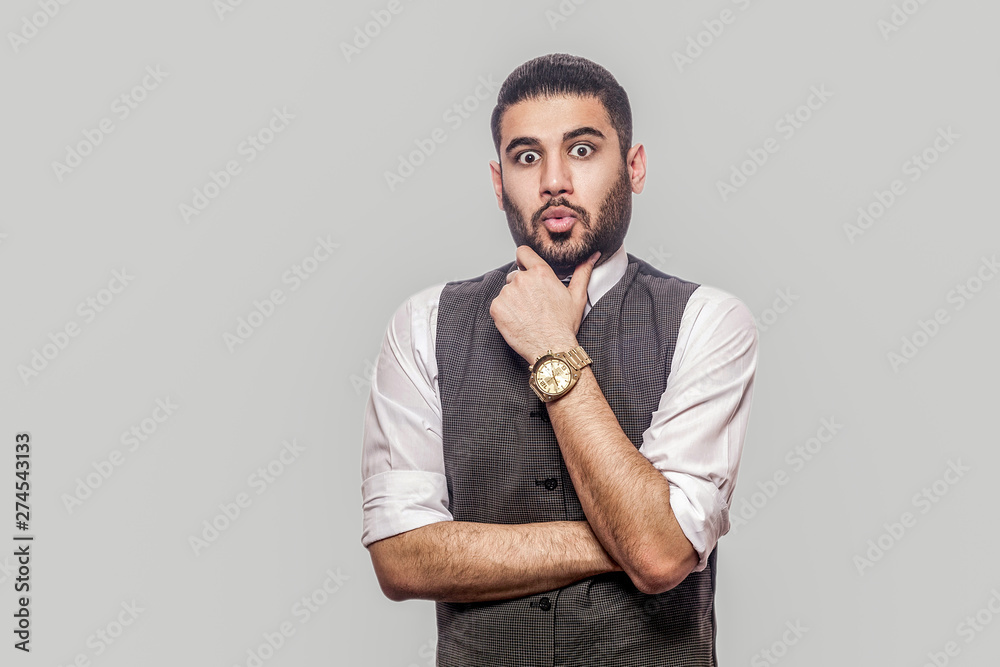 Portrait of suprised handsome bearded brunette man in white shirt and waistcoat standing, holding his chin and looking at camera with shocked face. indoor studio shot isolated on gray background. Portrait of suprised handsome bearded brunette man in white shirt and waistcoat standing, holding his chin and looking at camera with shocked face. indoor studio shot isolated on gray background.