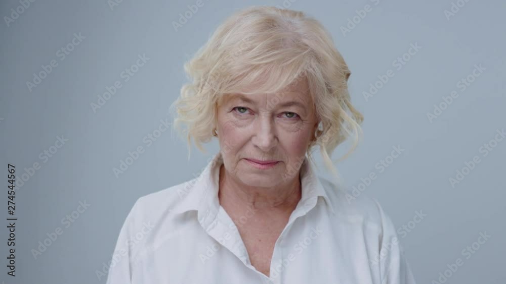 Attractive blonde mature woman in white shirt standing with serious expression against grey background looking on camera. Attractive blonde mature woman in white shirt standing with serious expression against grey background looking on camera.