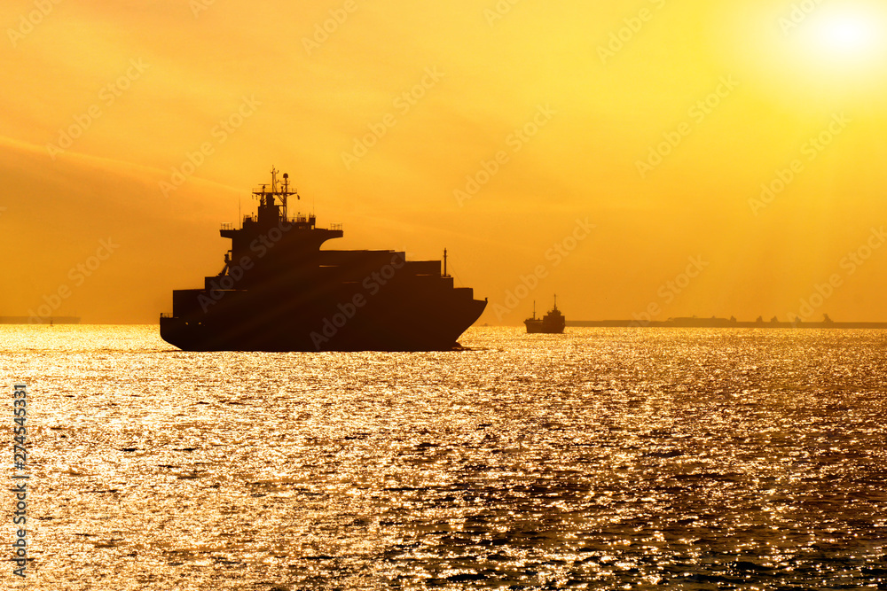 Silhouette of cargo ship with full shipload cargo containers are sailing in the Osaka bay evening with sun flare background.