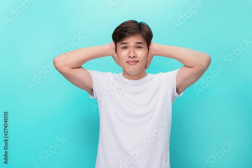 Too loud. Frustrated young man covering ears with hands and looking at camera while standing blue background