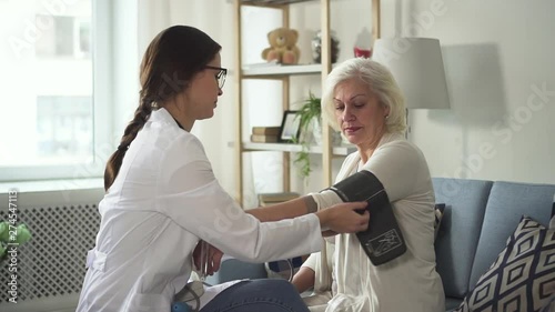 Professional young doctor assistant wearing in white coat uniform visit old lady patient in her cozy and charming flat with bright light room. Nurse measuring blood pressure using special equipment