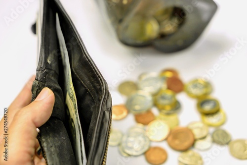 Hand holding black wallet with only one bank note inside and blurry coins open from the piggy bank laid out scattered on white background. This image signifies the use of money inexorably. photo