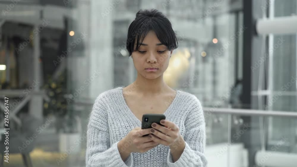 Middle shot of busy black hair asian female walking and holding scrolling a phone on background of glass windows in big mall center.