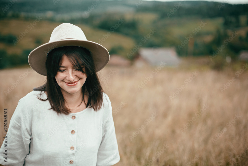 Stylish girl in linen dress and hat walking among herbs and wildflowers in field. Boho woman smiling and relaxing in countryside, simple slow life style. Space for text. Atmospheric image