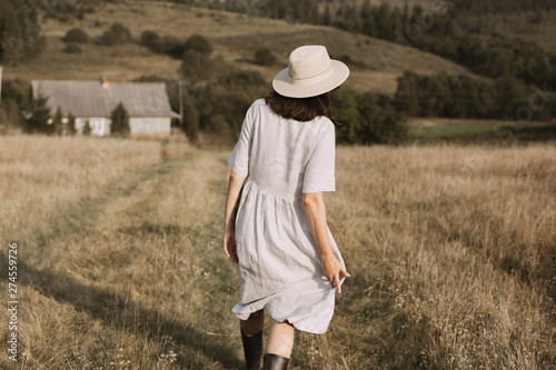Stylish girl in linen dress and hat walking among herbs and wildflowers in sunny field in mountains. Boho woman relaxing in countryside, simple rustic life. Atmospheric image. Space text © sonyachny